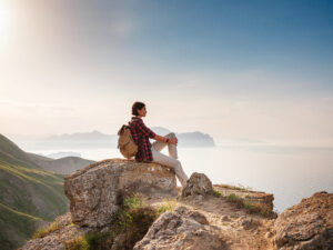 A young Asian woman with a backpack hiking in the summer sits on top of a montage and looks out over a beautiful sea bay landscape. Mountain and coastal travel, freedom and an active lifestyle