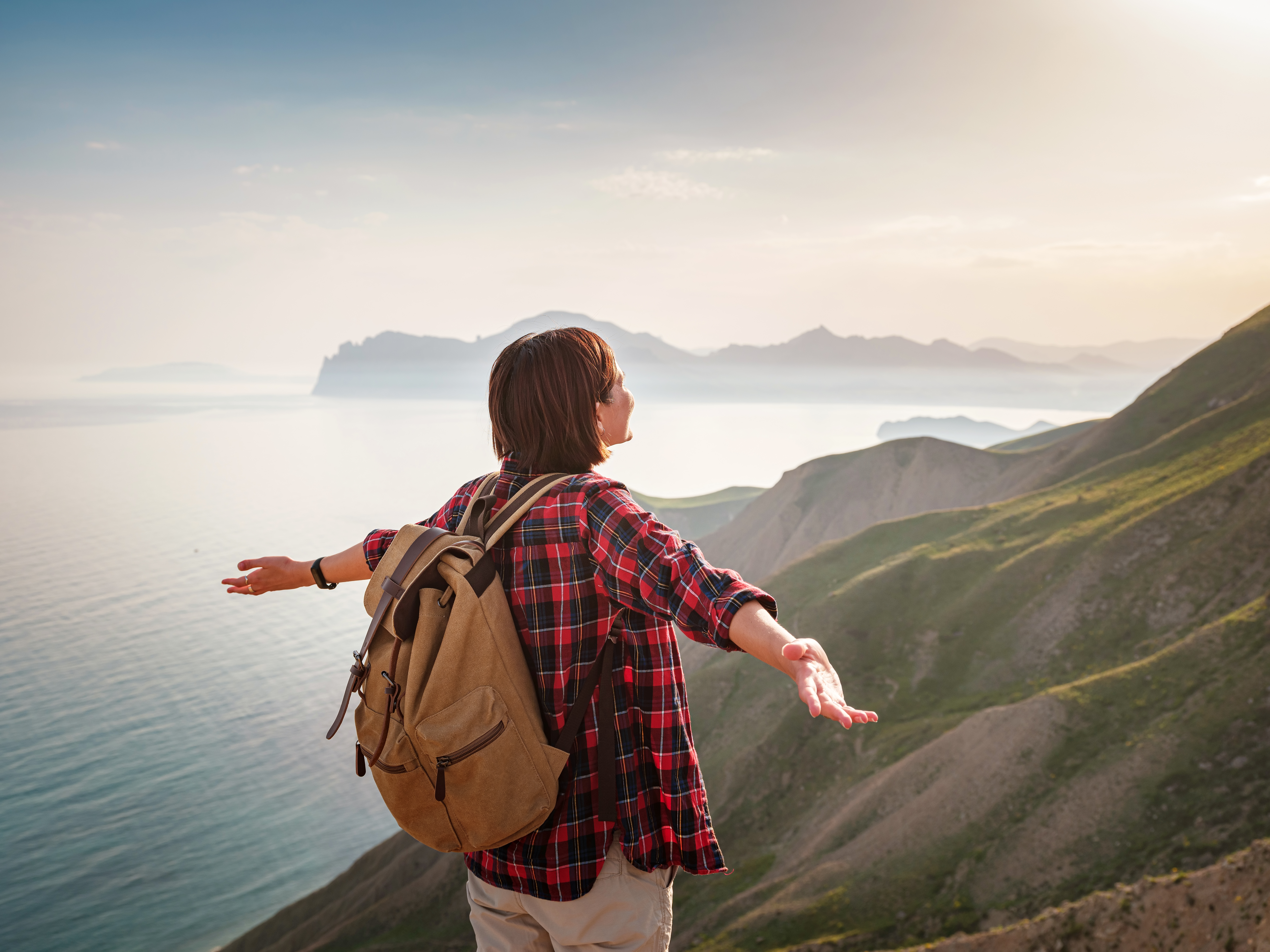 A young, happy woman with a backpack standing on a rock with her hands up and looking out over the sea below. Mountain and coastal travel, freedom and an active lifestyle
